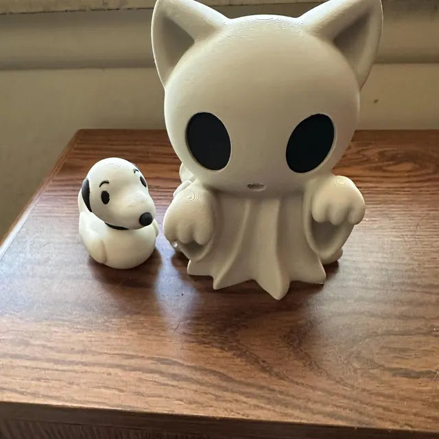 A ghost cat figurine with large black eyes and raised front paws is placed on a wooden table next to a small dog-shaped duck toy. The cat figure is designed in a ghostly white style with draped details resembling a sheet, creating a playful Halloween aesthetic. The smaller companion toy adds contrast and scale, making the pair an eye-catching decorative display for collectors and seasonal setups.
