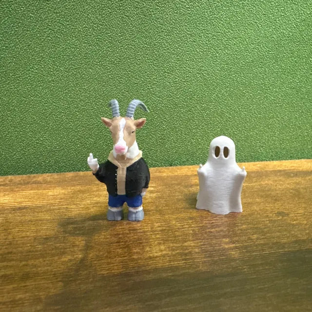 A small goat figurine dressed in a black jacket and blue pants stands with one hand raised in a playful gesture. Next to it is a simple white ghost figure with cut-out eyes, both placed on a wooden surface against a green background. The combination of the humorous goat character and the minimalist ghost decoration creates a quirky display suitable for collectors or seasonal décor setups.