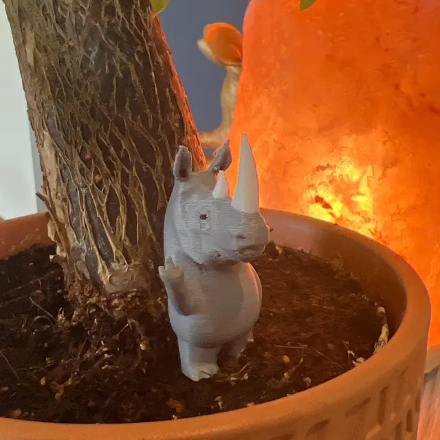 A small gray rhino figurine stands in the soil of a plant pot next to a tree trunk, positioned with one arm raised in a humorous gesture. The figure is highlighted by the warm glow of an orange lamp in the background, creating a cozy and playful atmosphere. Its compact size makes it a quirky decorative piece that adds personality to indoor plants or shelves.