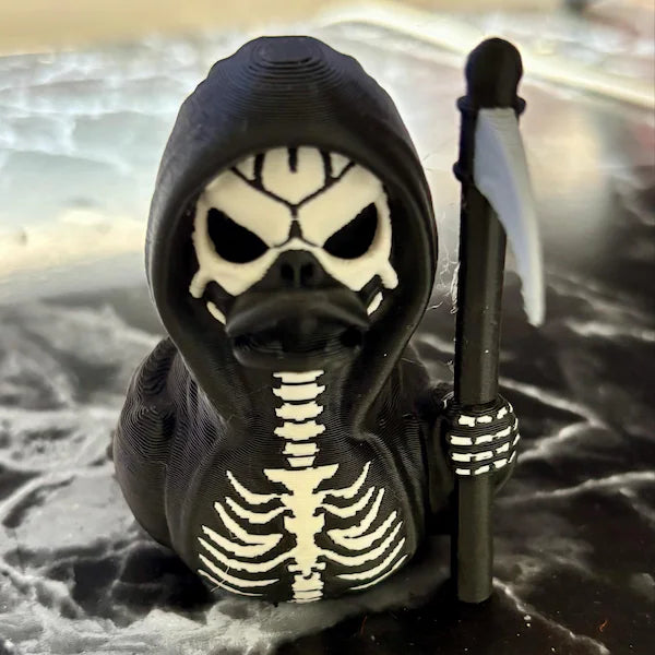 A Grim Reaper duck figurine sits on a marble-like surface, designed with black and white skeletal details, a dark hood, and a scythe in its wing. Its hollow eyes and bone-like patterns give it a spooky appearance, blending humor with a gothic Halloween theme. The figure’s compact size and playful design make it an ideal decorative piece or novelty collectible for those who enjoy eerie, creative accents in their seasonal displays.