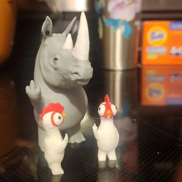 A gray rhino figurine stands upright with one arm raised in a playful gesture, positioned next to two small chicken figures with bright red crests and wide eyes. The combination of the larger rhino and the smaller chickens creates a humorous and whimsical scene. Displayed on a dark surface with household items in the background, the setup highlights the quirky and decorative appeal of these unique figurines.